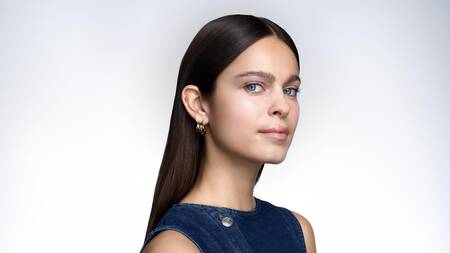 Portrait d'une femme aux cheveux fonces, yeux bleus, boucles d'oreilles dorees et haut en jean. Regard direct.